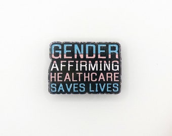 Peut inclure: Épinglette en émail noir avec le texte "Gender Affirming Healthcare Saves Lives" en lettres blanches. Le texte "Gender" est en rayures bleues et roses.