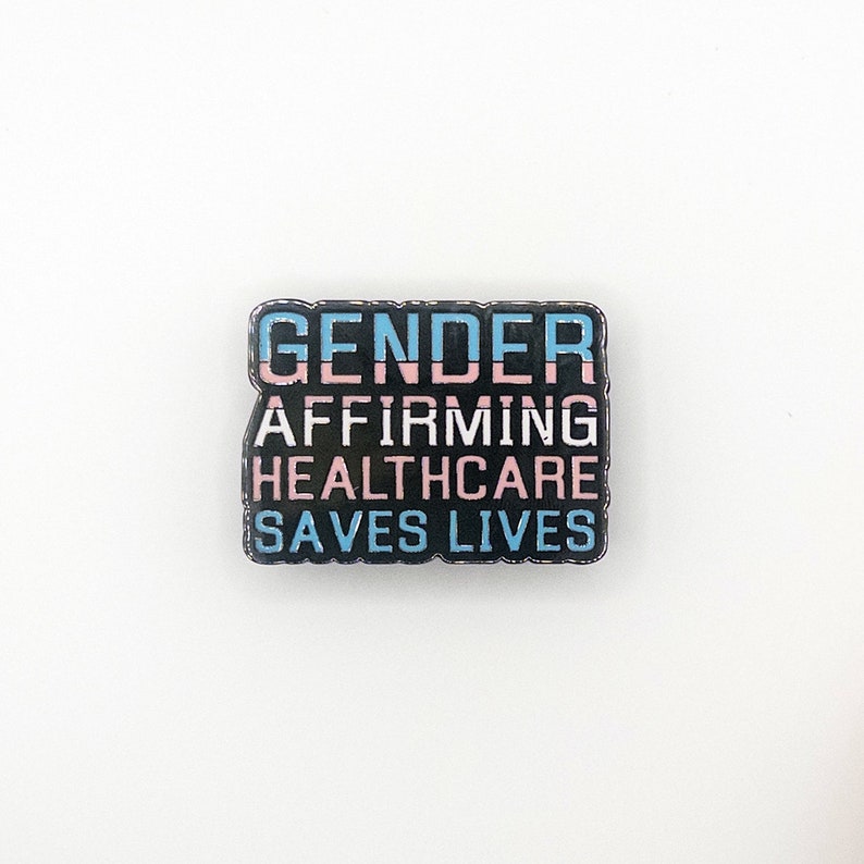May include: Black enamel pin with the text "Gender Affirming Healthcare Saves Lives" in white lettering. The text "Gender" is in blue and pink stripes.