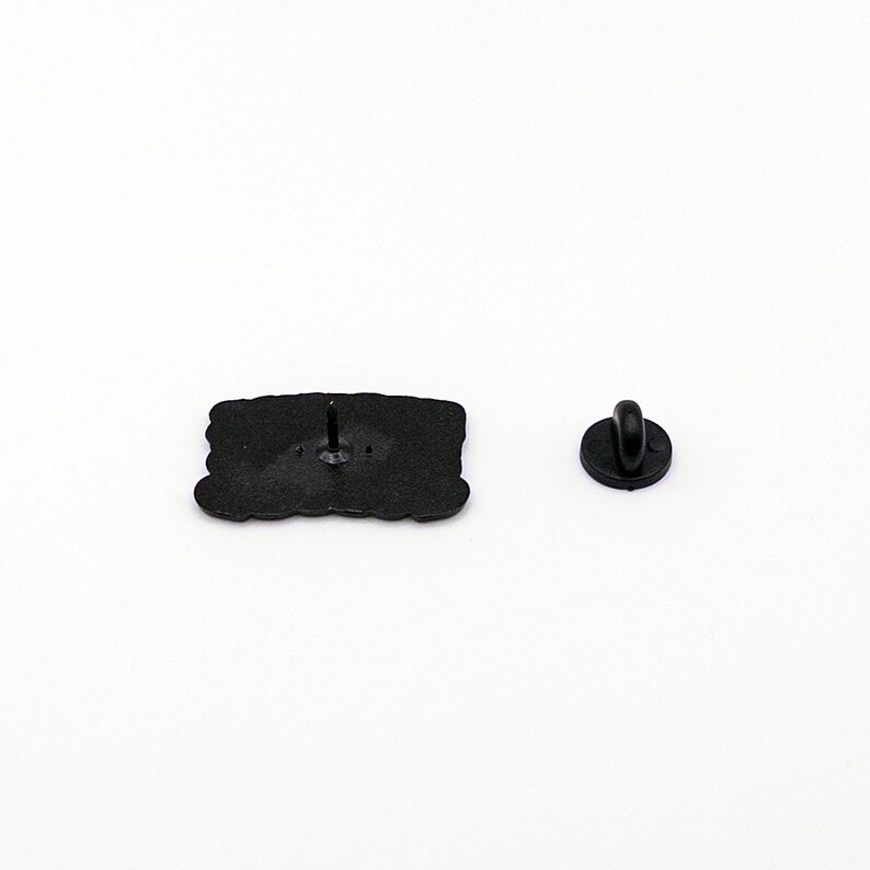 May include: Black enamel pin featuring a cartoon character with a speech bubble. The pin has a metal back with a butterfly clasp.