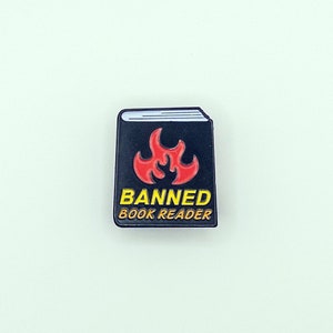 May include: Black enamel pin shaped like a book with a red flame and the text "Banned Book Reader".