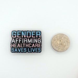 May include: A black enamel pin with the text "GENDER AFFIRMING HEALTHCARE SAVES LIVES" in white letters. The text is outlined in blue and pink, resembling the transgender pride flag.
