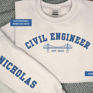 May include: Sand-colored sweatshirt with blue text that reads "CIVIL ENGINEER" above a bridge graphic and "EST. 2025." The sleeve displays "NICHOLAS" in blue. The sweatshirt is customizable with name and date.