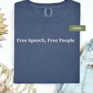 May include: A denim blue t-shirt with the text "Free Speech, Free People" in white. The shirt is folded neatly, with a tag visible at the neckline. A pair of blue jeans is partially visible in the background.