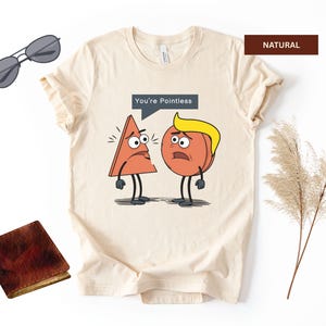 May include: Cream-colored t-shirt with a cartoon graphic of a triangle and a peach with the text "You're Pointless". The shirt also has a pair of sunglasses and a small brown wallet.