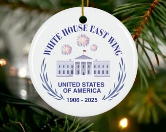 May include: A white, round Christmas ornament with the text "WHITE HOUSE EAST WING" in a circular pattern. The ornament features an illustration of the White House, fireworks, and the text "UNITED STATES OF AMERICA 1906-2025".