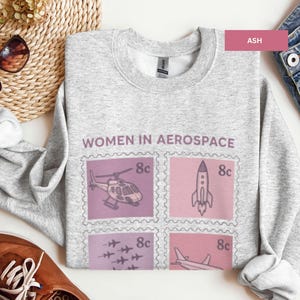 May include: Light gray sweatshirt with the text "WOMEN IN AEROSPACE" and postage stamp-style graphics of a helicopter, rocket, jets, and airplane. The sweatshirt is ash-colored and has a crew neck.
