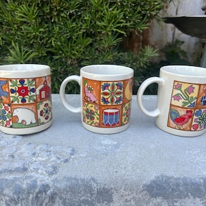 Op de afbeelding: Drie keramische mokken met witte handvatten en een crèmekleurige basis. Elke mok heeft een kleurrijk patchwork-ontwerp met verschillende illustraties, waaronder een haan, bloemen en een beer. De mokken zijn gerangschikt op een grijs oppervlak.