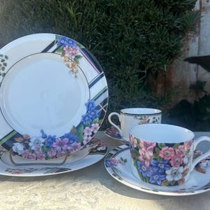 May include: A set of floral patterned teacups and saucers. The cups and saucers feature a black and white striped border with a floral design in shades of pink, blue, and green.