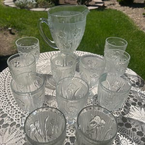 May include: A set of clear glass pitcher and glasses with a floral design. The pitcher and glasses have a textured surface and are arranged on a white lace tablecloth.
