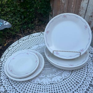 May include: A stack of white ceramic plates with a delicate pink floral pattern and a light blue border. The plates are arranged on a white lace tablecloth, with additional plates stacked nearby. The plates are a classic dinnerware set.