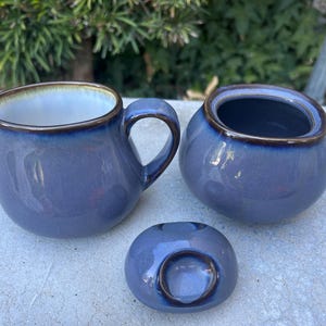 May include: A three-piece ceramic set in a muted blue-gray glaze. The set includes a creamer with a handle, a sugar bowl, and a lid. Each piece has a contrasting brown rim.