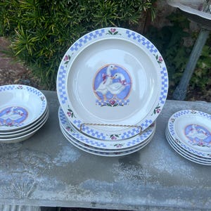 Puede incluir: Un juego de vajilla de cerámica blanca con un borde a cuadros azul y blanco. Cada pieza tiene un diseño central de dos gansos con lazos rosas. El juego incluye cuencos, platos llanos y platos más pequeños.