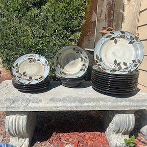 May include: A set of three ceramic plates with a leaf design in shades of brown, green, and blue. The plates are stacked on a concrete bench with red mulch in the background.