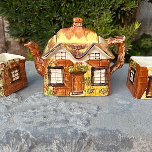 May include: A brown and white ceramic teapot and two matching cups shaped like a cottage with a thatched roof. The teapot has a spout and handle, and the cups have handles. The cottage has windows and a door.
