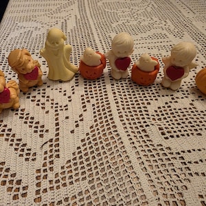 May include: A collection of Halloween-themed figurines, including a ghost, cherubs with hearts, pumpkins, and dogs with hearts. The figurines are in shades of orange, yellow, and white, arranged on a white crocheted tablecloth.