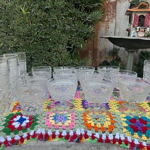 May include: A collection of vintage clear glass drinkware, including tall tumblers, stemmed dessert dishes, and smaller glasses, arranged on a colorful, patterned crochet blanket. The glasses have a diamond pattern.