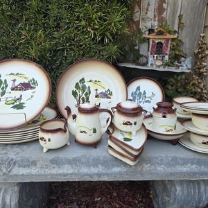 May include: A collection of cream-colored ceramic dinnerware featuring a brown rim and a green and brown farm scene illustration. The set includes plates, a teapot, a sugar bowl, a creamer, small dishes, and cups with matching saucers.