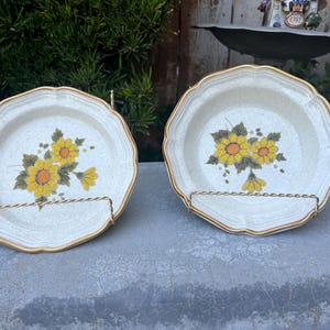May include: Two white ceramic bowls with a gold rim and a floral design featuring yellow flowers and green leaves. The bowls are on a gold metal stand.