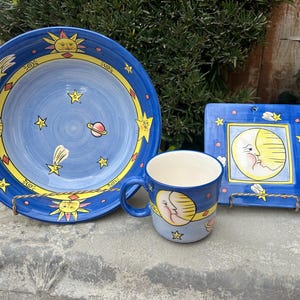 May include: A blue and yellow ceramic set of a bowl, mug, and small plate with a celestial theme. The designs feature a sun, moon, stars, and planets.