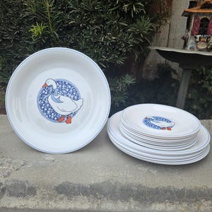 May include: White ceramic plates with a blue rim and a goose illustration. The goose is in a blue floral circle with a blue bow. The plates are stacked in two piles, one large plate and a stack of smaller plates.