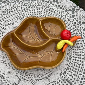 May include: A brown ceramic serving dish shaped like a leaf with three sections. The dish has a raised design of veins on the leaf. There are three small ceramic vegetables on the dish: a red pepper, a yellow pepper, and a carrot.