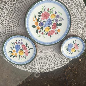 Könnte beinhalten: Set aus drei Keramiktellern mit einem Blumendesign mit Trauben, Pfirsichen und Blumen in den Farben Rosa, Blau, Gelb und Grün. Die Teller haben einen blauen und weißen Rand mit einem grünen Rand.