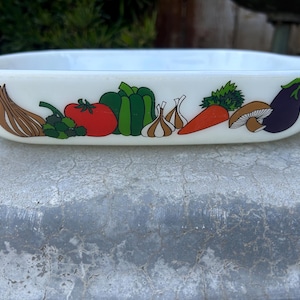 May include: A rectangular, off-white casserole dish with a colorful vegetable design. The design includes illustrations of a tomato, bell pepper, garlic, carrot, mushroom, and eggplant. The dish is likely vintage.