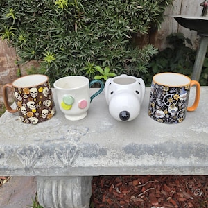 May include: Four ceramic mugs with unique designs. One mug features skulls, another has macaron-like decorations, a third is shaped like a dog's head, and the last has a floral and gnome pattern. Each mug has a different colored handle.