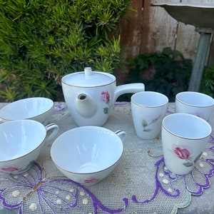 May include: A white tea set with a teapot and five cups. The teapot and cups are decorated with pink roses and green leaves. The set is on a white lace tablecloth with a purple butterfly design.