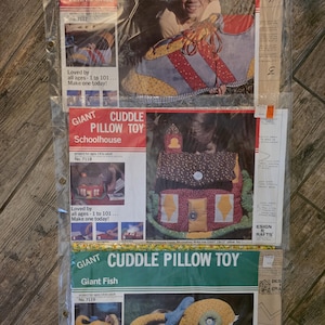 May include: Three craft project kits for giant cuddle pillow toys, including a tennis shoe, a schoolhouse, and a fish. Each kit is packaged in a clear plastic sleeve with printed instructions, photographs of the finished product, and the text Giant Cuddle Pillow Toy.
