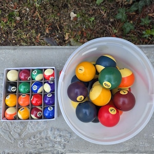 May include: A set of billiard balls, some in a white box and others in a clear plastic container. The balls are various colors, including red, yellow, green, and blue, with white stripes and numbers.
