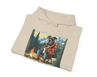 May include: A beige hoodie featuring a graphic of a dog wearing a plaid shirt and sitting in a chair by a campfire in a snowy mountain scene. The dog is holding a mug. The text "Camp Dog" is visible at the bottom of the image.