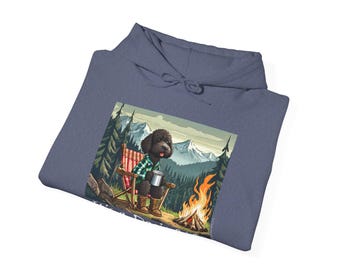 May include: A dark blue hoodie featuring a graphic of a black dog sitting in a camping chair by a campfire in a woodland setting. The dog is wearing a plaid shirt and holding a mug. The text "Camp Life" is printed below the graphic.
