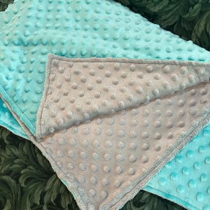 May include: A close-up of two plush blankets. One is a light turquoise color, and the other is a light gray color. Both blankets have a textured, dotted surface. The gray blanket is folded over the turquoise blanket.
