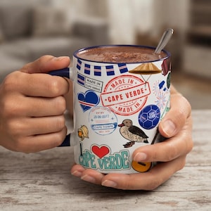 Op de afbeelding: Een witte keramische mok met een donkerblauw handvat en rand, voorzien van kleurrijke reisthema-afbeeldingen waaronder een rode stempel met de tekst MADE IN CAPE VERDE, een blauw hart, een vogelillustratie, een voetbal en de tekst I LOVE CAPE VERDE.