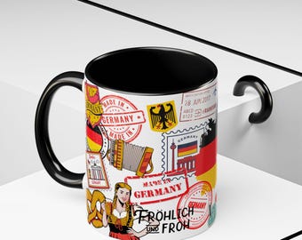 May include: A white ceramic mug with a black interior and handle, decorated with German-themed stamps, flags, and a woman in traditional attire. The mug features the text "Made in Germany" and "Fröhlich und Froh."