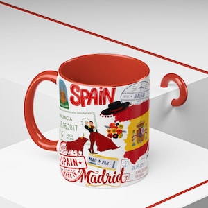 May include: A ceramic mug with a red interior, handle, and rim. The exterior features a design with the word "SPAIN" in red, along with illustrations of a matador, a bull, and the Spanish flag. The mug has travel-themed stamps and text.