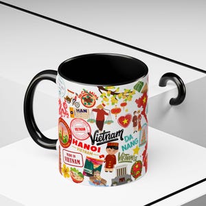 Pode incluir: Uma caneca de cerâmica branca com interior e alça pretos. A caneca é decorada com ilustrações coloridas e texto relacionados ao Vietname, incluindo as palavras "Vietnam", "Hanoi" e "Da Nang".