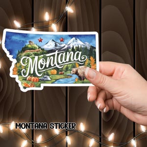 May include: A Montana sticker shaped like the state, with a watercolour illustration of mountains, a river, a bear, and pumpkins. The word "Montana" is written in cursive. The sticker has the text "MONTANA STICKER" at the bottom.