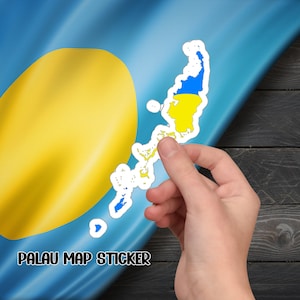May include: A hand holding a sticker of the Palau islands. The sticker is blue, yellow, and white and is shaped like the islands. The sticker is on a wooden background. The text "PALAU MAP STICKER" is visible below the sticker.