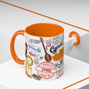Pode incluir: Caneca de cerâmica branca com interior e alça laranja. Decorada com selos, ilustrações e texto relacionados ao Chipre, incluindo um mapa, um instrumento musical e a frase "Made in Cyprus".