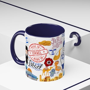 Pode incluir: Caneca de cerâmica branca com interior e alça azul marinho. A caneca é decorada com várias ilustrações de temática israelense, incluindo um selo que diz "Made in Israel", um camelo e uma flor vermelha.