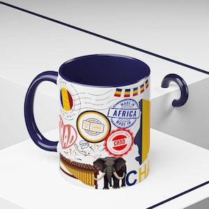 May include: A white ceramic mug with a navy blue interior and handle. The mug is decorated with stamps, flags, and illustrations related to Chad, including an elephant and a xylophone. The text "Made in Africa" is also visible.