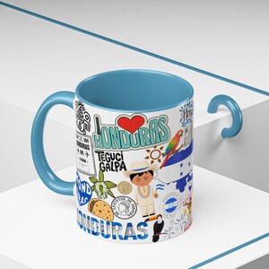 May include: A white ceramic mug with a blue interior and handle, featuring a colorful design with the word "Honduras", a red heart, and illustrations of Honduran culture, including a toucan and a map.