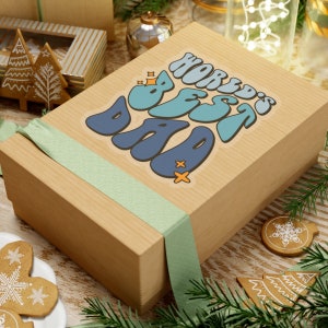 K&ouml;nnte beinhalten: Eine braune Geschenkbox mit einem gr&uuml;nen Band und einem Aufkleber mit der Aufschrift "World's Best Dad" in blauen, gr&uuml;nen und orangen Buchstaben.