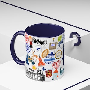 May include: A white ceramic mug with a navy blue interior and handle. The mug is decorated with colorful illustrations and the word "Finland" in various designs, including stamps, landmarks, and cultural symbols.
