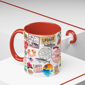 Puede incluir: Una taza de cerámica blanca con interior y asa rojos, decorada con sellos e ilustraciones con temática de Florida. El diseño incluye imágenes de palmeras, una matrícula, un tiburón y el texto "FLORIDA".