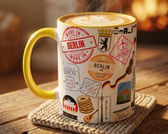May include: A white ceramic mug with a yellow handle, filled with coffee and latte art. The mug is decorated with travel-themed stamps and the text "MADE IN BERLIN". It sits on a beige knitted coaster.