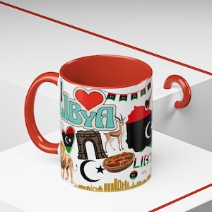 May include: A white ceramic mug with a red interior, handle, and rim. The mug features various Libyan-themed graphics, including a map, flags, and the word "Libya" in multiple places. The design includes a heart, a gazelle, and a crescent moon.