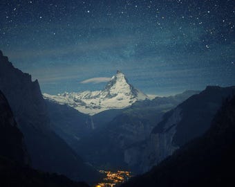 Può includere: Una cima montuosa innevata si erge contro un cielo blu scuro punteggiato di stelle. La montagna è circondata da colline boscose scure.